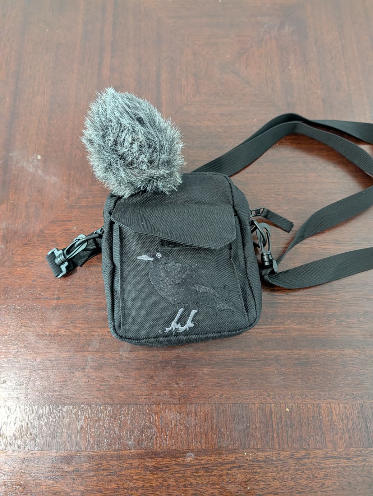A small black shoulder bag with an embroidered crow, a furry windshield poking out of the top